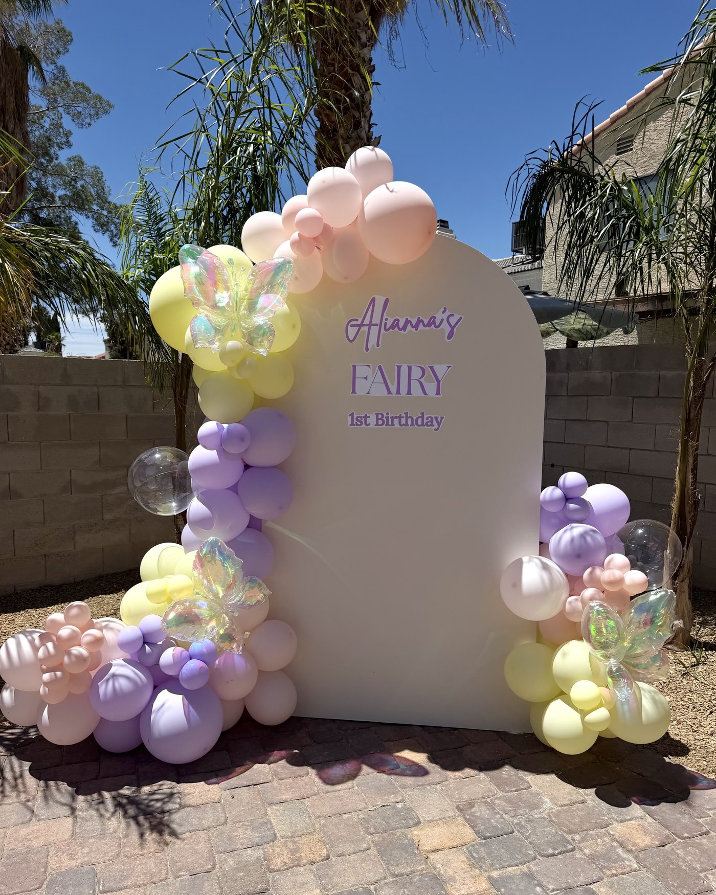 A pastel arched balloon backdrop for a first birthday, with lavender, yellow and pink balloons and iridescent butterflies